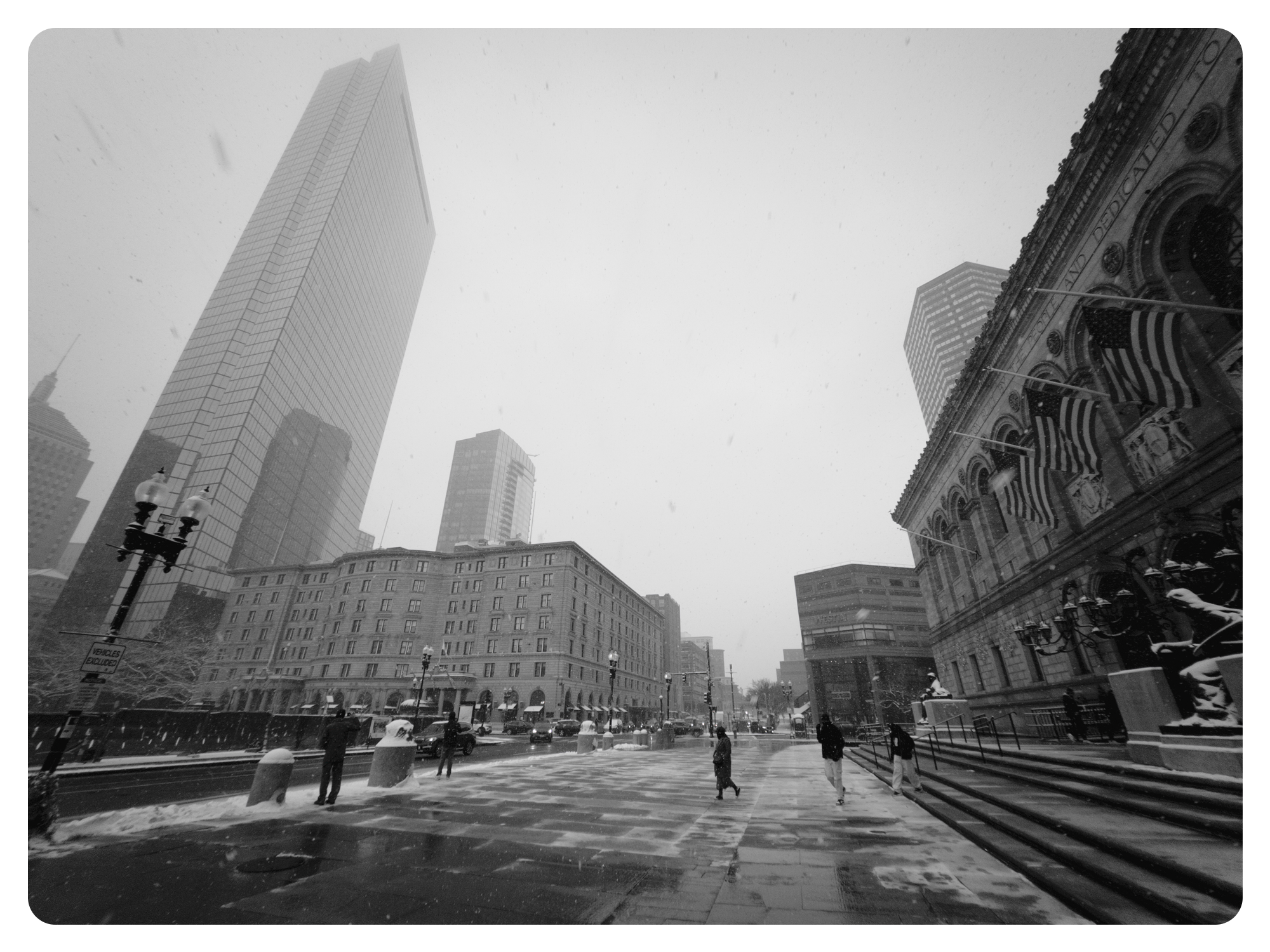 B&W ultrawide photo of Copley Square in Boston. The sky is overcast with snow visibly falling in the frame. pictured on the left is 230 Clarendon, and on the right is Boston Public Library. snow lightly coats parts of the sidewalks and steps up to the library. 